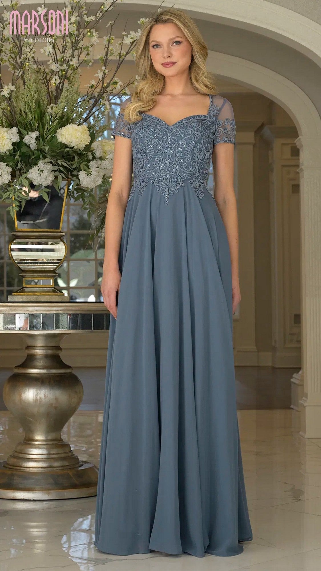 A woman stands indoors wearing the Marsoni M271, a long formal slate blue dress with a chiffon pleated skirt, short sheer sleeves, a sweetheart neckline, and intricate embroidery on the bodice.